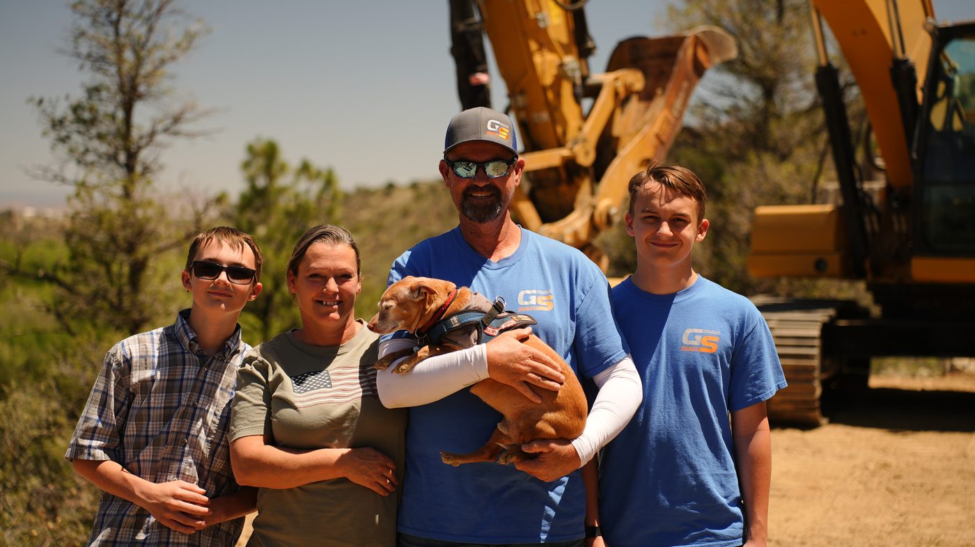 The Grading Specialists family standing together in front of a CAT excavator on a Yavapai County job site